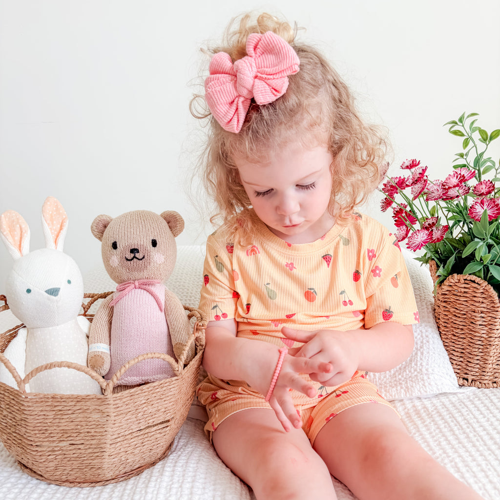 Child wearing a yellow outfit with fruit and heart patterns, sitting on a bed with toys and flowers in the background.