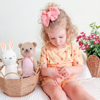 Child wearing a yellow outfit with fruit and heart patterns, sitting on a bed with toys and flowers in the background.