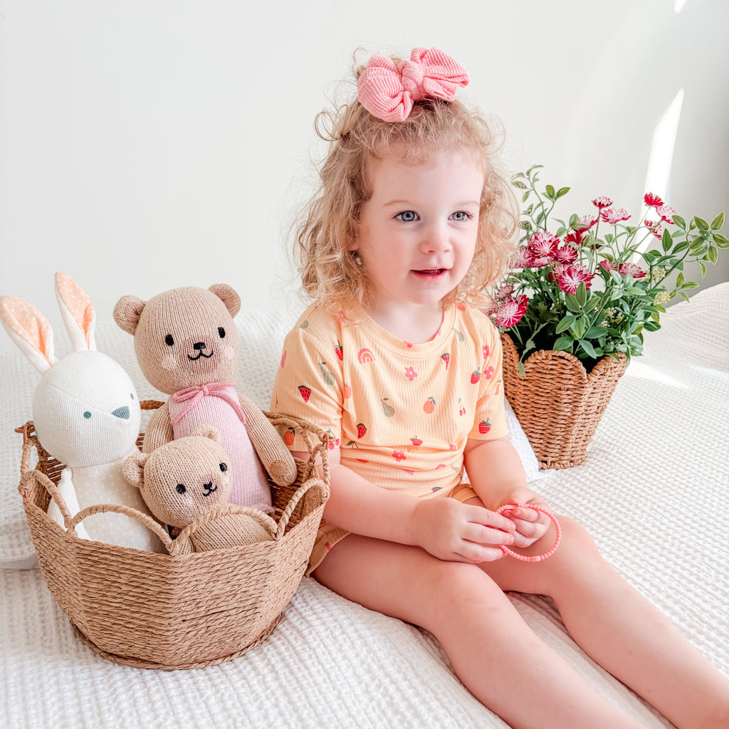 Child wearing a yellow outfit with fruit and heart patterns, sitting on a bed with toys and flowers in the background.