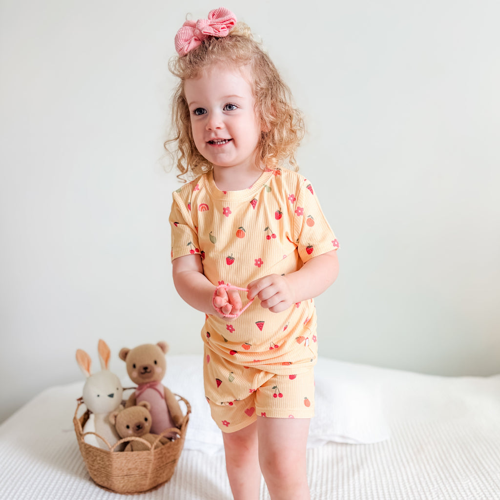 Child in a yellow outfit with fruits & hearts patterns standing on a bed with stuffed toys in the background.