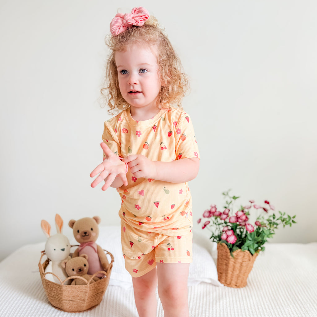 Child in a yellow outfit with heart and fruit patterns standing on a bed with toys and plants.