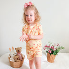 Child in a yellow outfit with heart and fruit patterns standing on a bed with toys and plants.