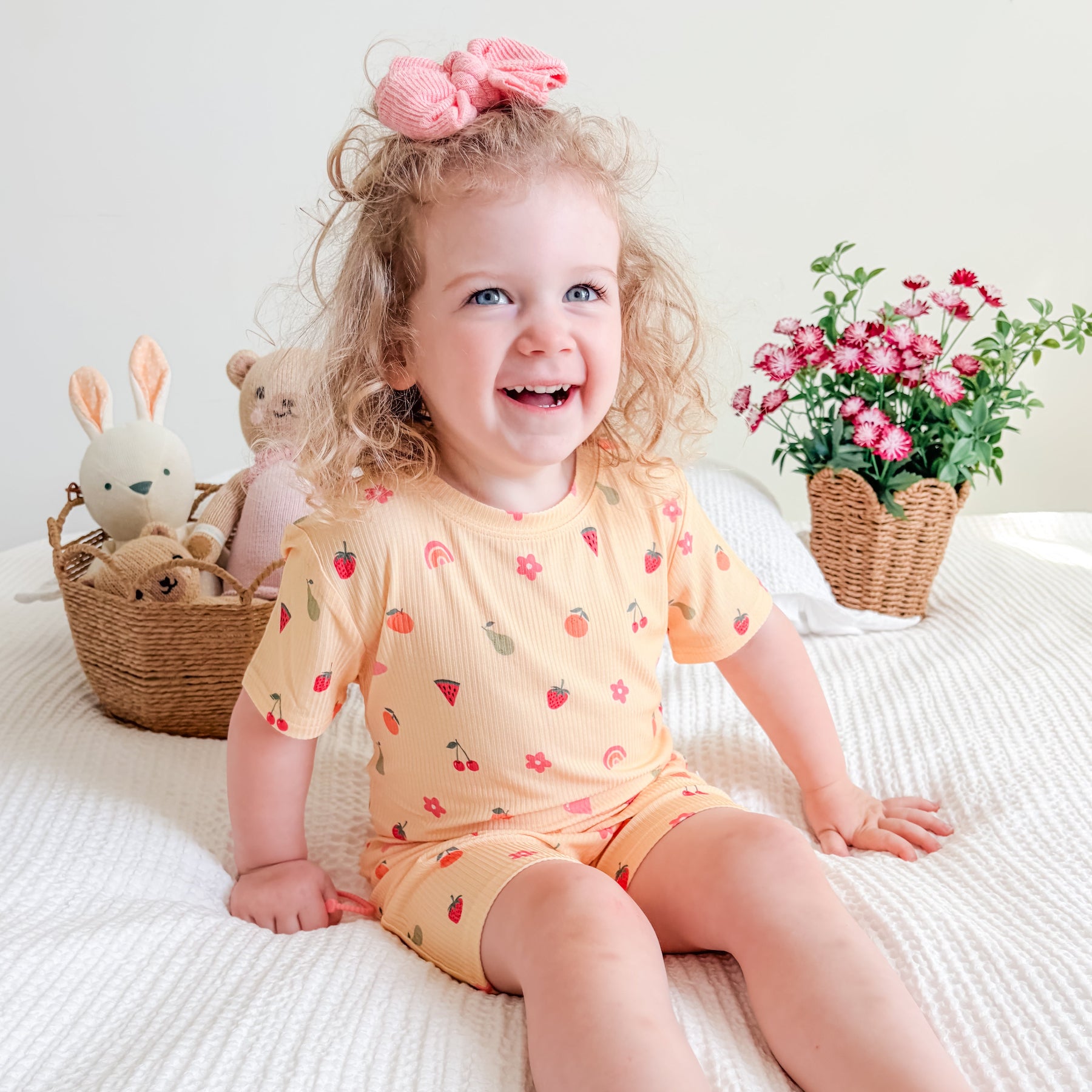 Child wearing a yellow outfit with fruit and heart patterns, sitting on a bed with toys and flowers in the background.