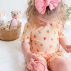Child wearing a yellow outfit with fruit and heart patterns, sitting on a bed with toys in the background.