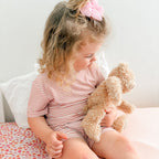 Child sitting on a bed holding a teddy bear, wearing a pink and white striped shorts pajama set.