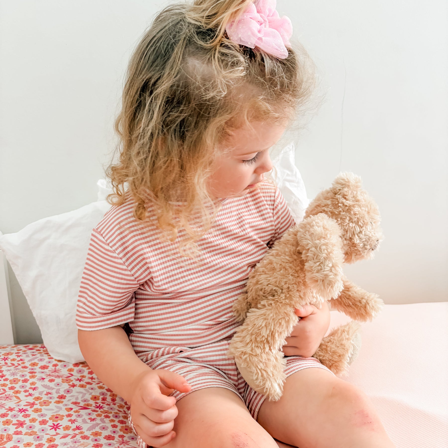 Child sitting on a bed holding a teddy bear, wearing a pink and white striped shorts pajama set.