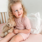 Child sitting on a bed holding a teddy bear, wearing a pink and white striped shorts pajama set.