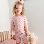 Child wearing a pink and white striped shorts pajamas holding a teddy bear in a bedroom