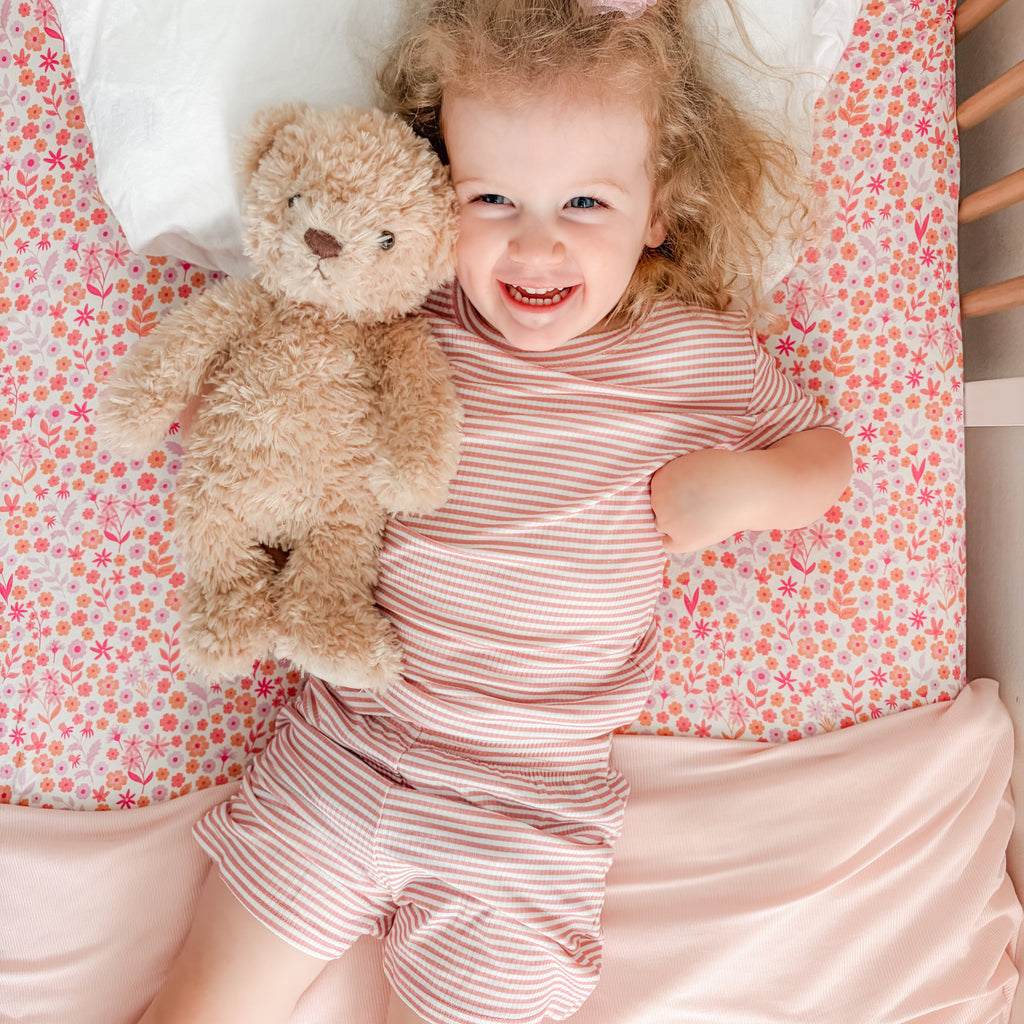 Child in a pink striped outfit holding a teddy bear on a floral bedspread