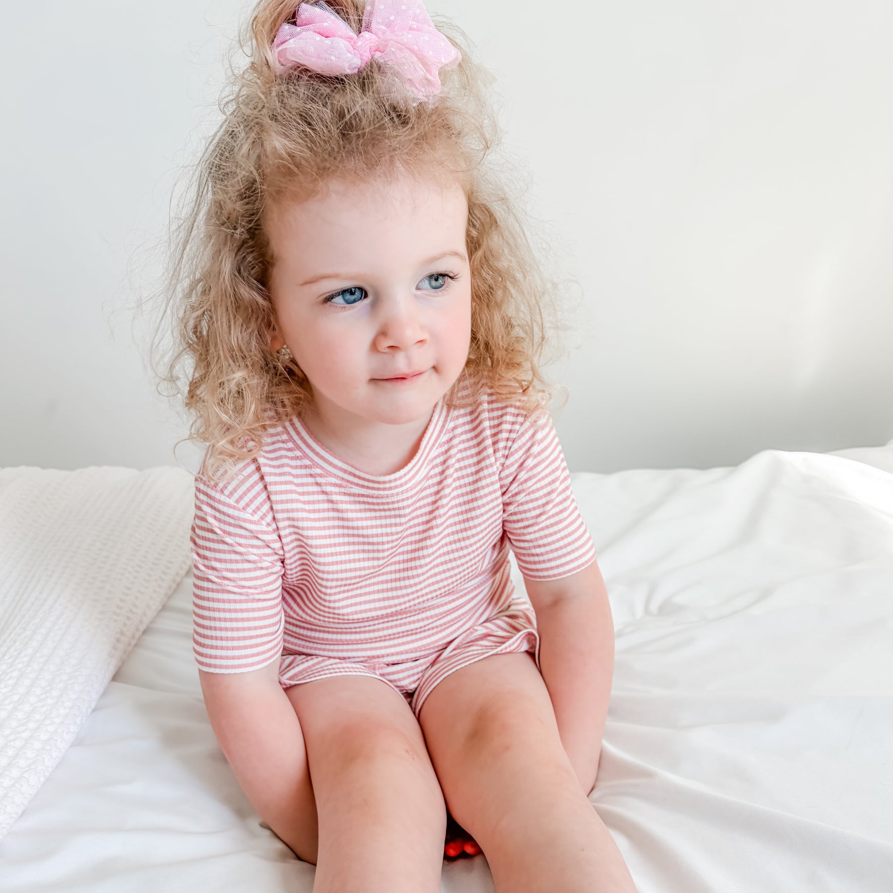 Child sitting on a bed wearing a pink and white striped shorts pajama set.