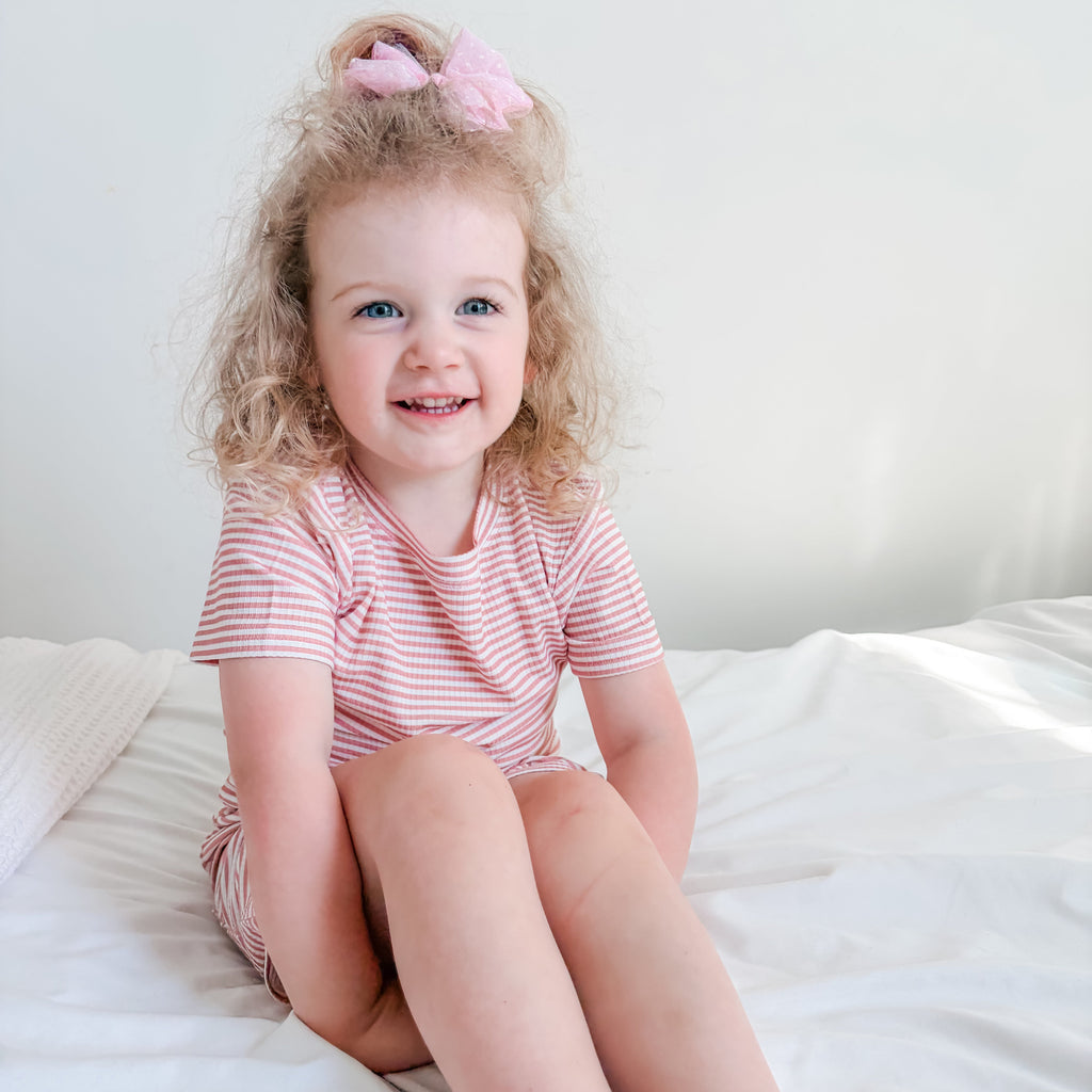 Child sitting on a bed wearing a pink and white striped shorts pajama set.