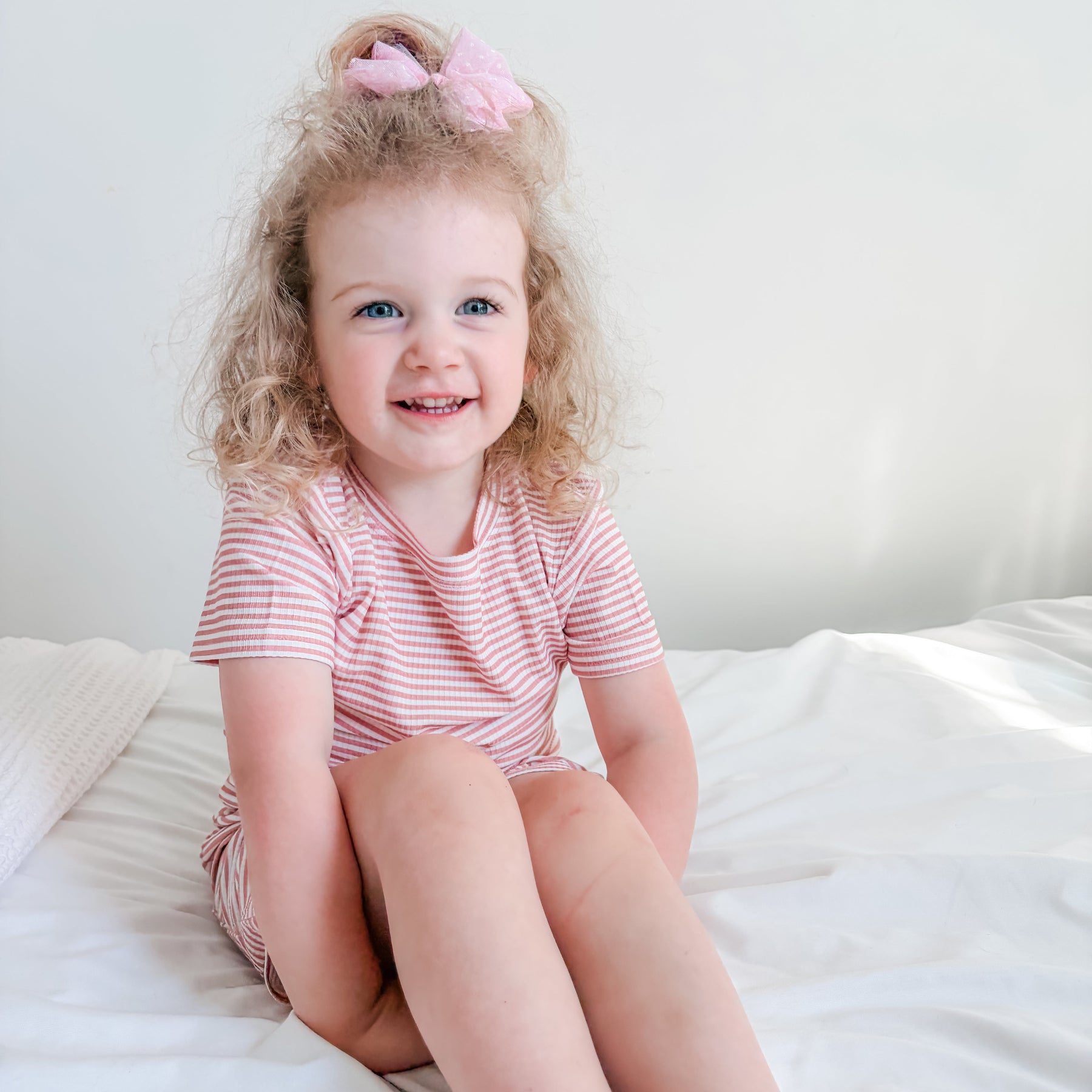 Child sitting on a bed wearing a pink and white striped shorts pajama set.