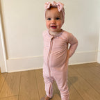 Baby in a pink striped outfit with a headband standing on a wooden floor.