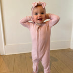 Baby in a pink and white striped onesie with a matching headband standing on a wooden floor.