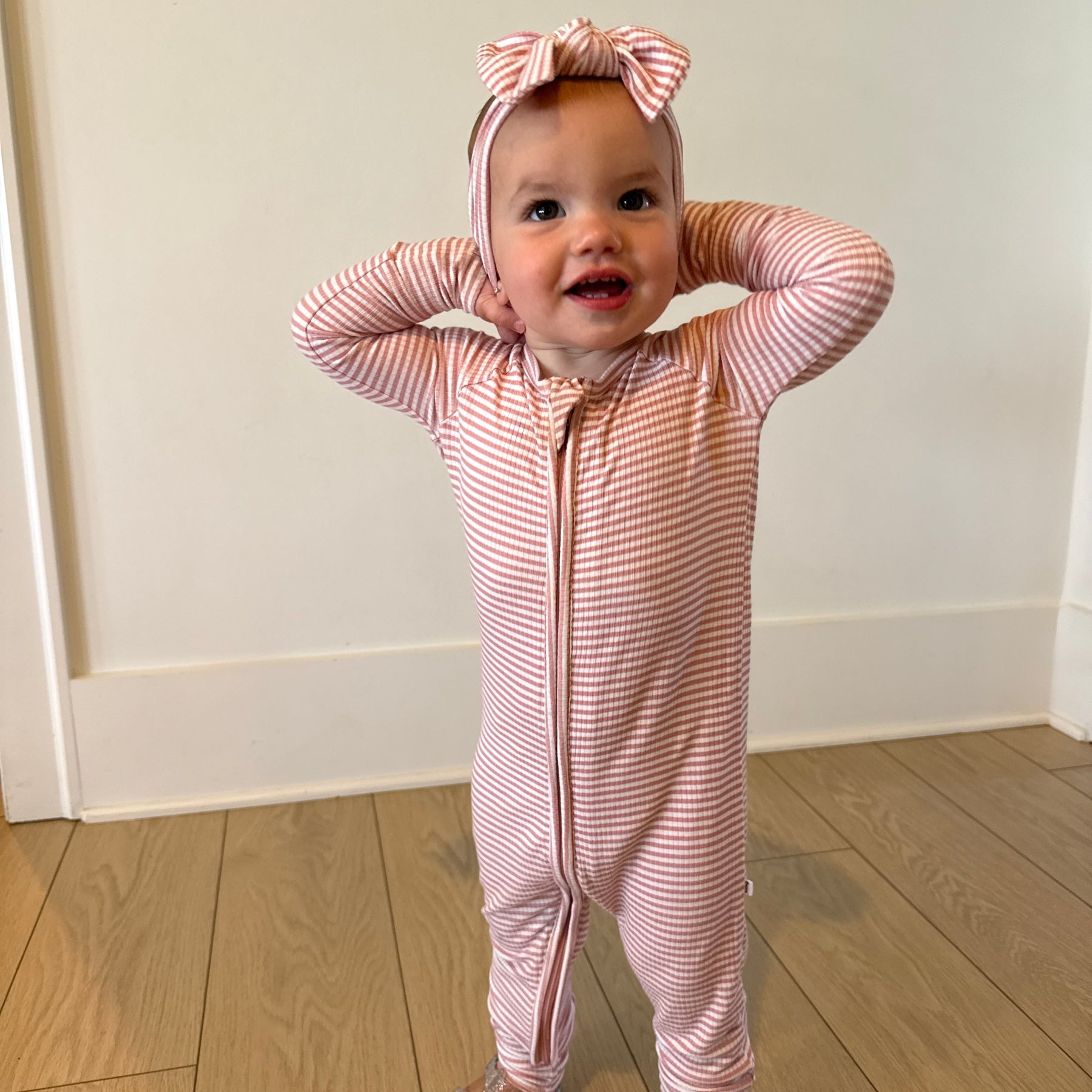 Baby in a pink and white striped onesie with a matching headband standing on a wooden floor.