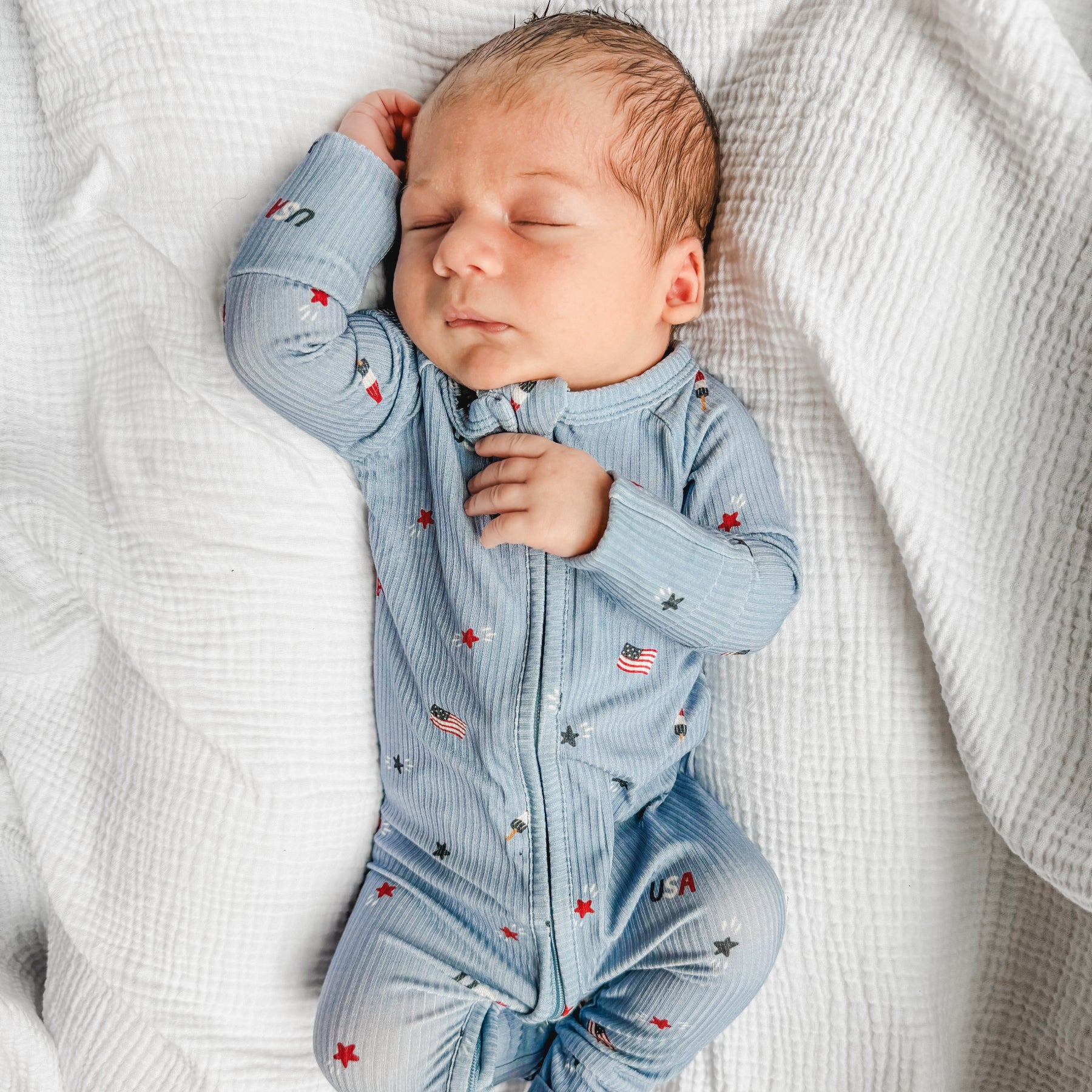 Newborn baby sleeping in a blue onesie with stars and flags on a white textured background.