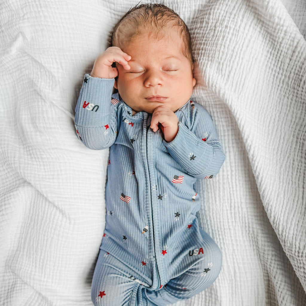Newborn baby sleeping in a blue onesie with stars and flags on a white textured background.