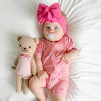 Baby in pink shortie with teddy bear and pacifier, wearing a large pink bow headband.