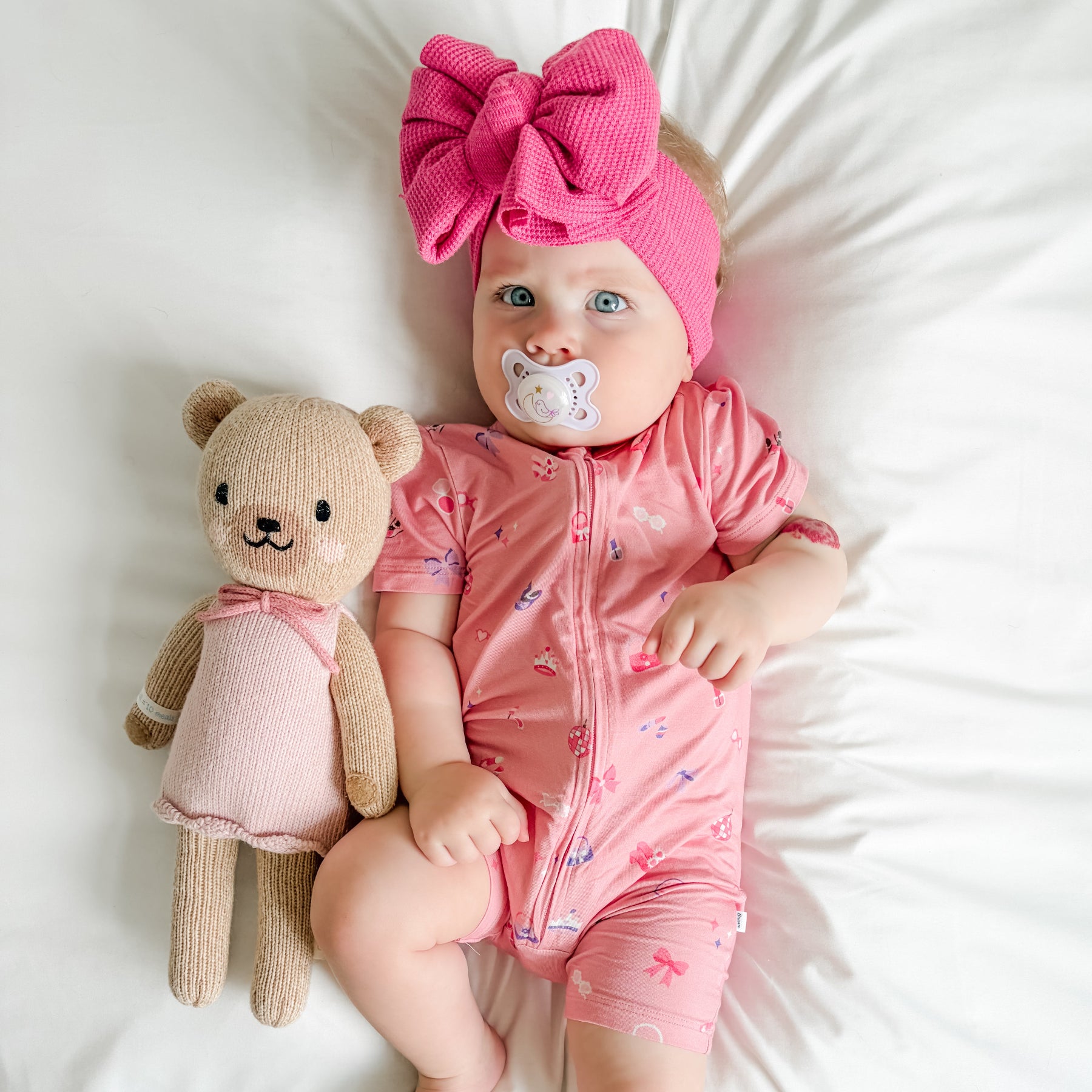 Baby in pink shortie with teddy bear and pacifier, wearing a large pink bow headband.