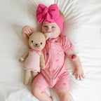 Baby in pink shortie with teddy bear and pacifier, wearing a large pink bow headband.