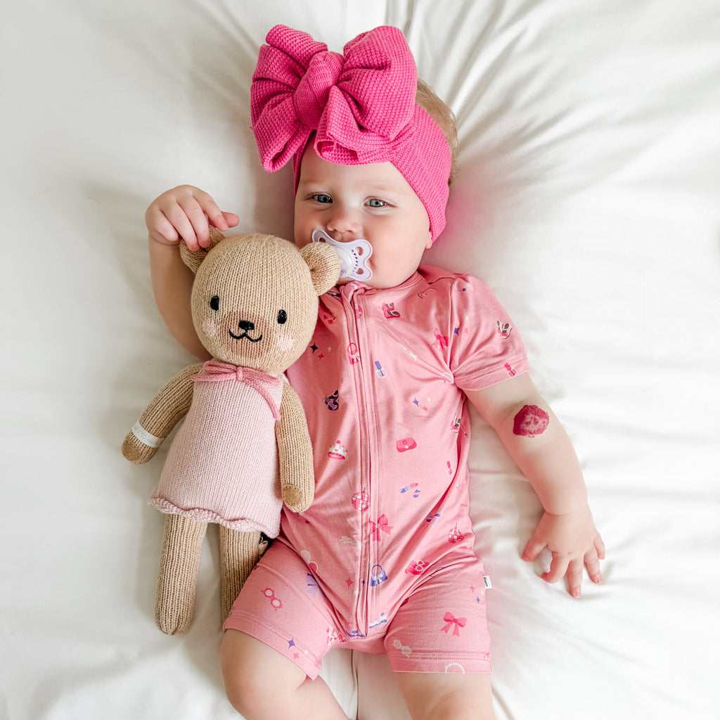 Baby in pink shortie with teddy bear and pacifier, wearing a large pink bow headband.
