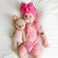 Baby in pink shortie with teddy bear and pacifier, wearing a large pink bow headband.