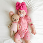 Baby in pink shortie with teddy bear and pacifier, wearing a large pink bow headband.