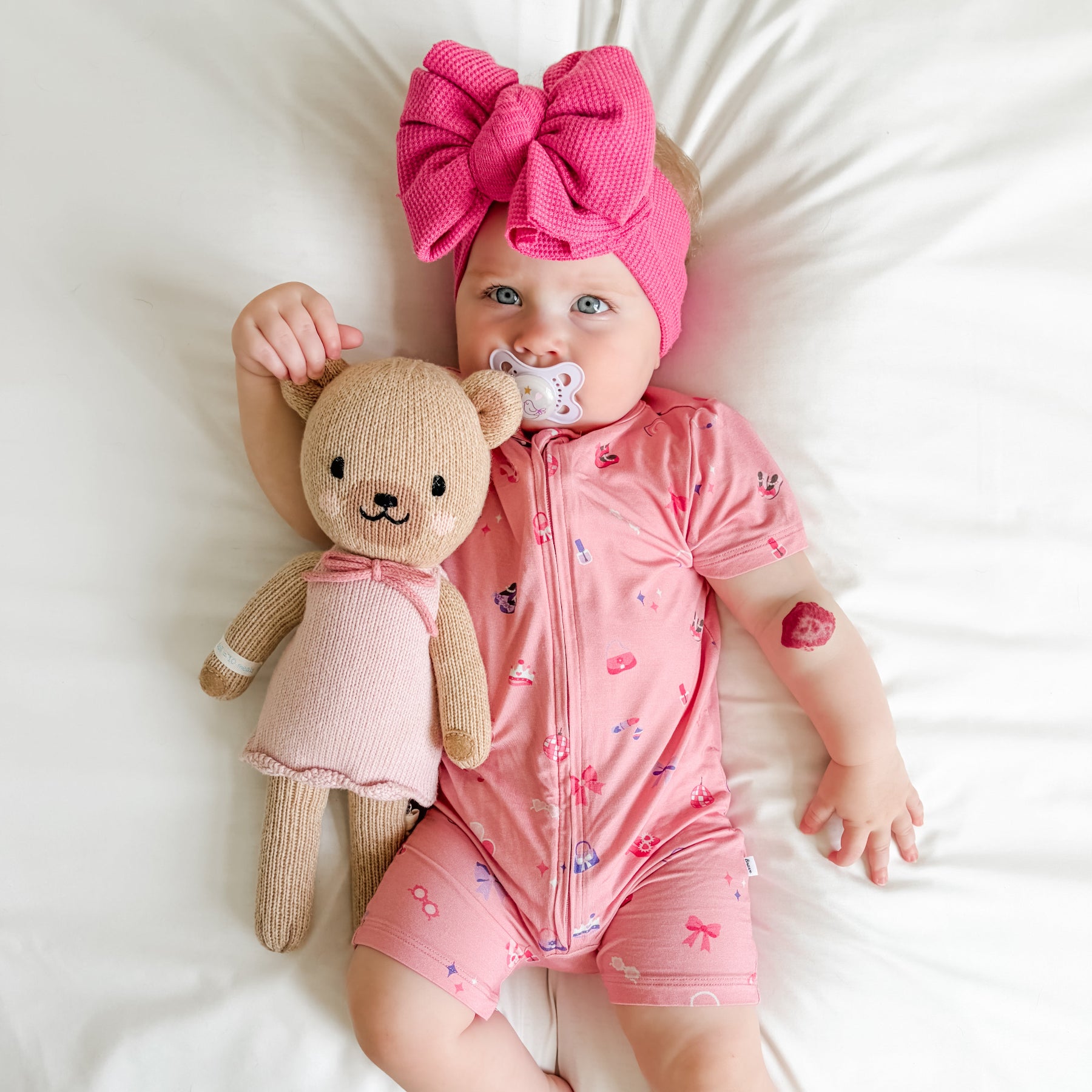 Baby in pink shortie with teddy bear and pacifier, wearing a large pink bow headband.