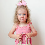 Young girl in a pink dress with a bow in her hair, holding a small bag against a plain background