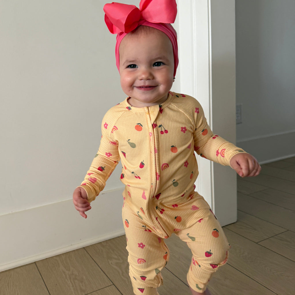 Baby in a yellow onesie with fruit patterns and a pink bow headband standing on a wooden floor.