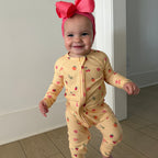 Baby in a yellow onesie with fruit patterns and a pink bow headband standing on a wooden floor.