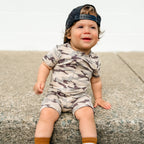 Child wearing a camouflage onesie and cap sitting on a concrete surface.