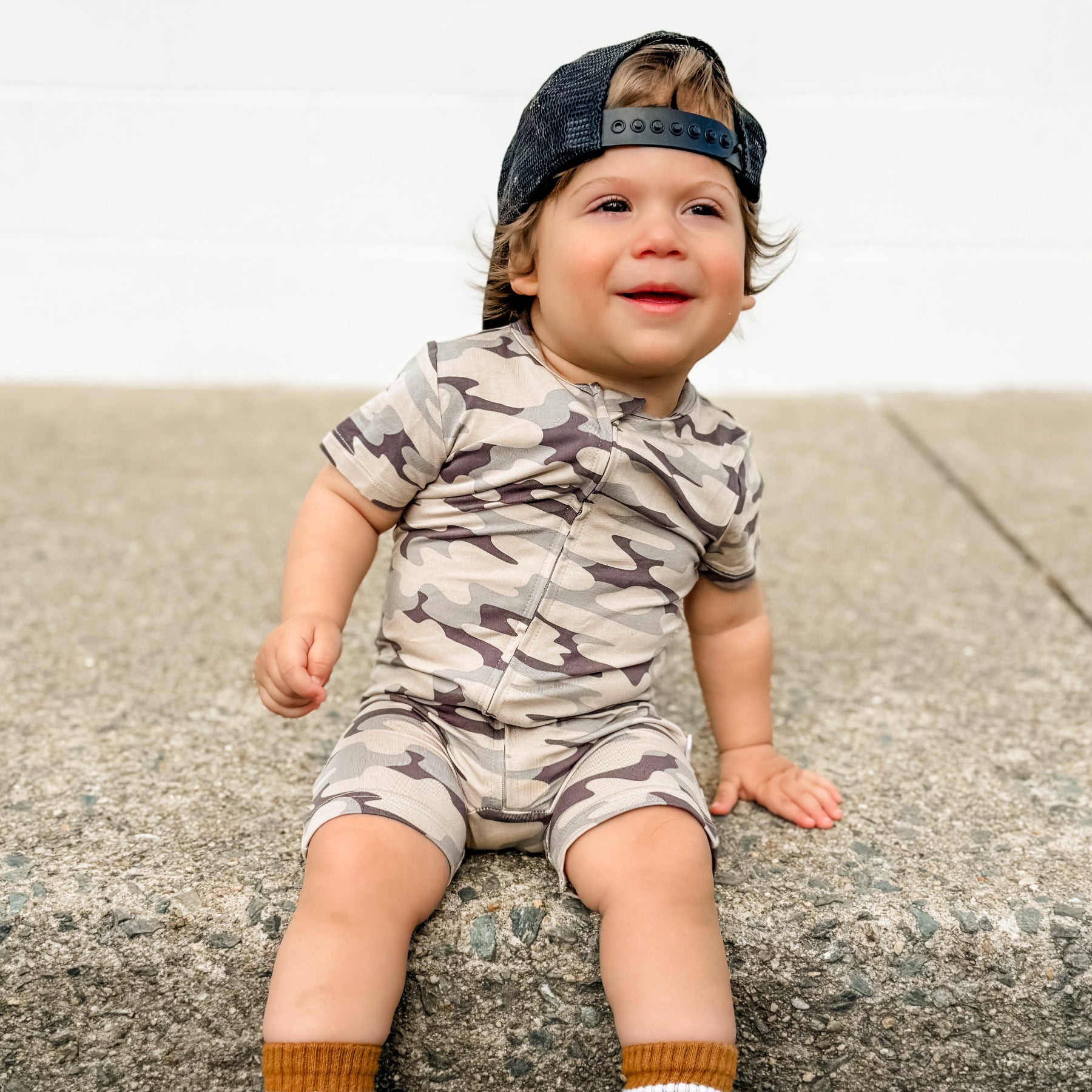 Child wearing a camouflage onesie and cap sitting on a concrete surface.