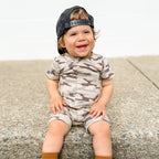 Child wearing a camouflage onesie and cap sitting on a concrete surface.