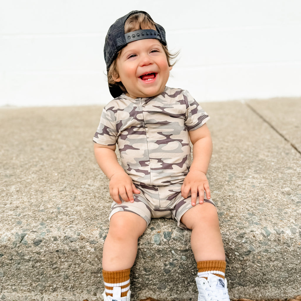 Child wearing a camouflage onesie and cap sitting on a concrete surface.