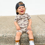 Child wearing a camouflage onesie and cap sitting on a concrete surface.