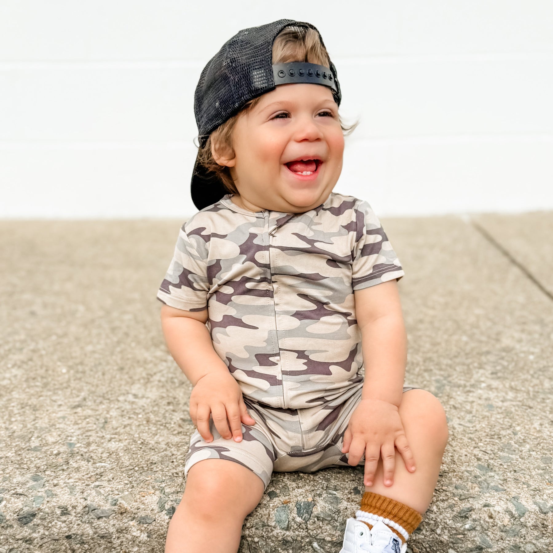 Child wearing a camouflage onesie and cap sitting on a concrete surface.