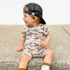 Child wearing a camouflage onesie and cap sitting on a concrete surface.