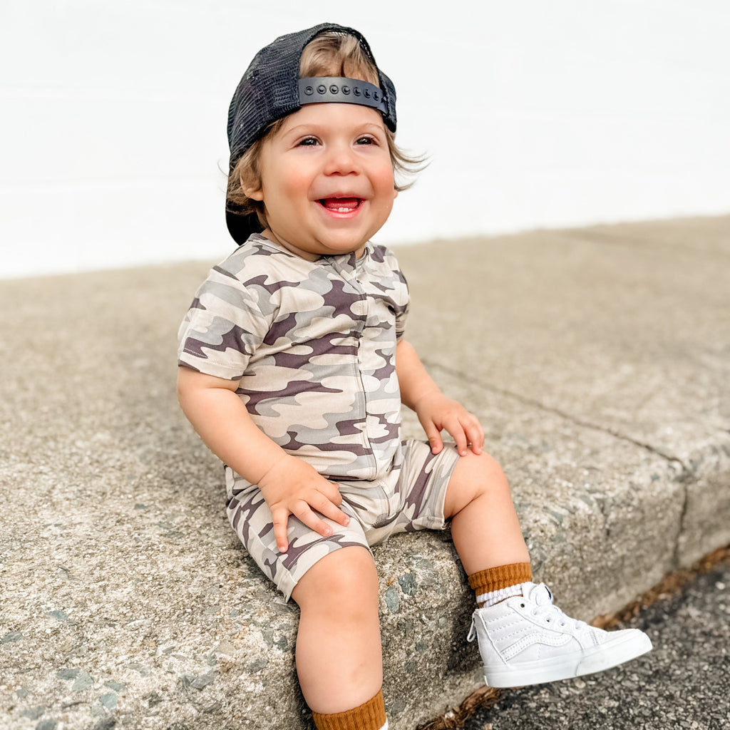 Child wearing a camouflage onesie and cap sitting on a concrete surface.