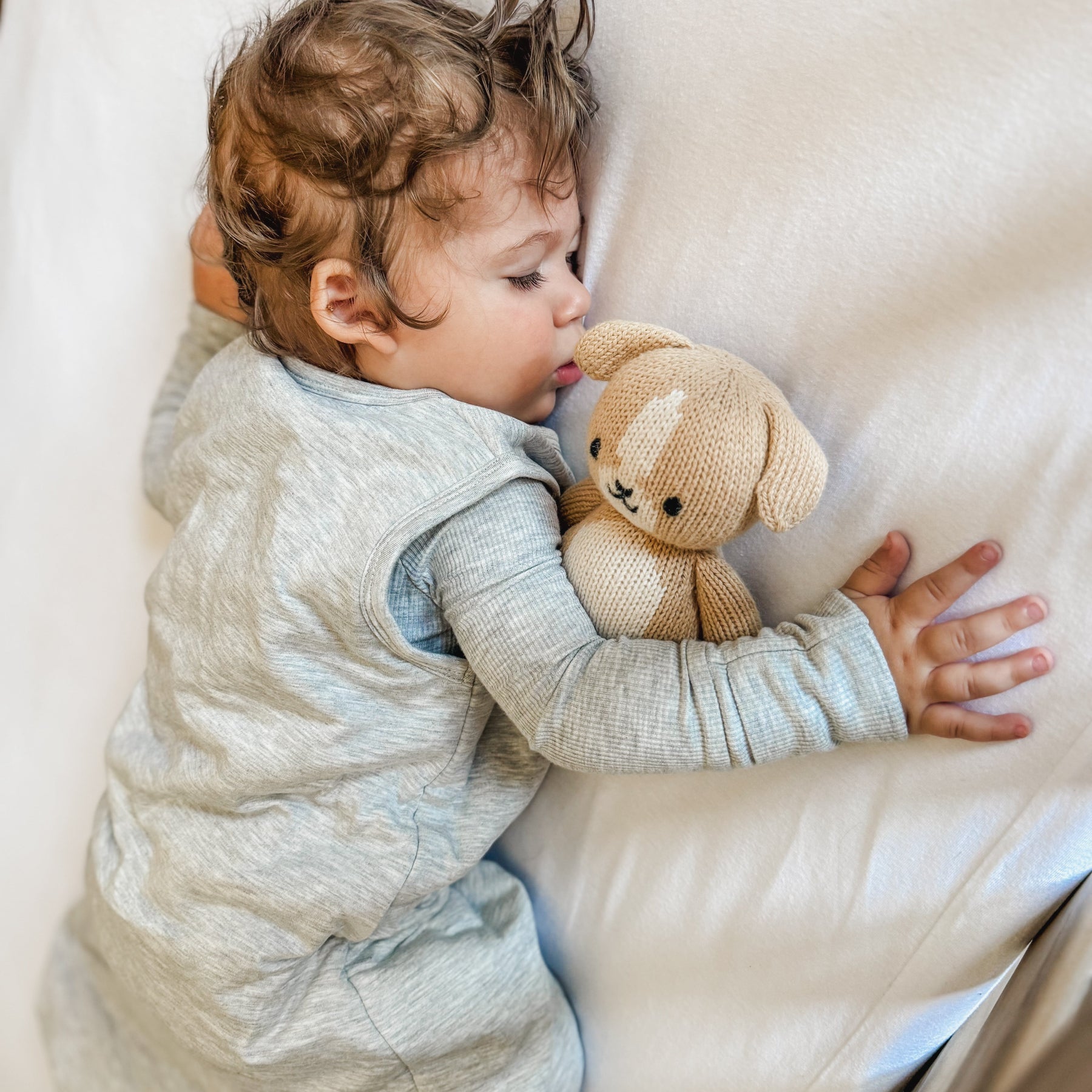 Child sleeping in a crib with white bedding, wearing a gray sleeping bag, holding a teddy bear