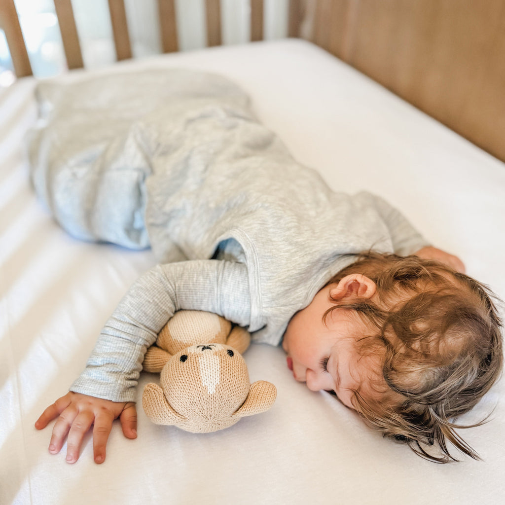 Child sleeping in a crib with white bedding, wearing a gray sleeping bag, holding a teddy bear