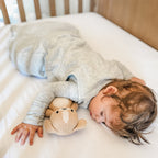 Child sleeping in a crib with white bedding, wearing a gray sleeping bag, holding a teddy bear