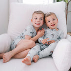Two children in matching pajamas sitting on a white chair with a plant in the background.