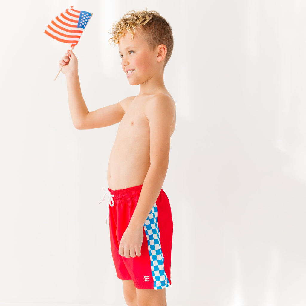 Young boy holding an American flag and wearing red swim trunks with a checkered pattern on a white background