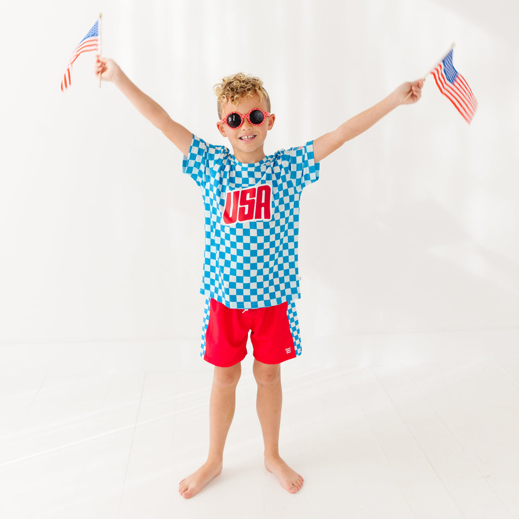 Child wearing a blue 'USA' t-shirt with checkered shorts, holding American flags on a white background
