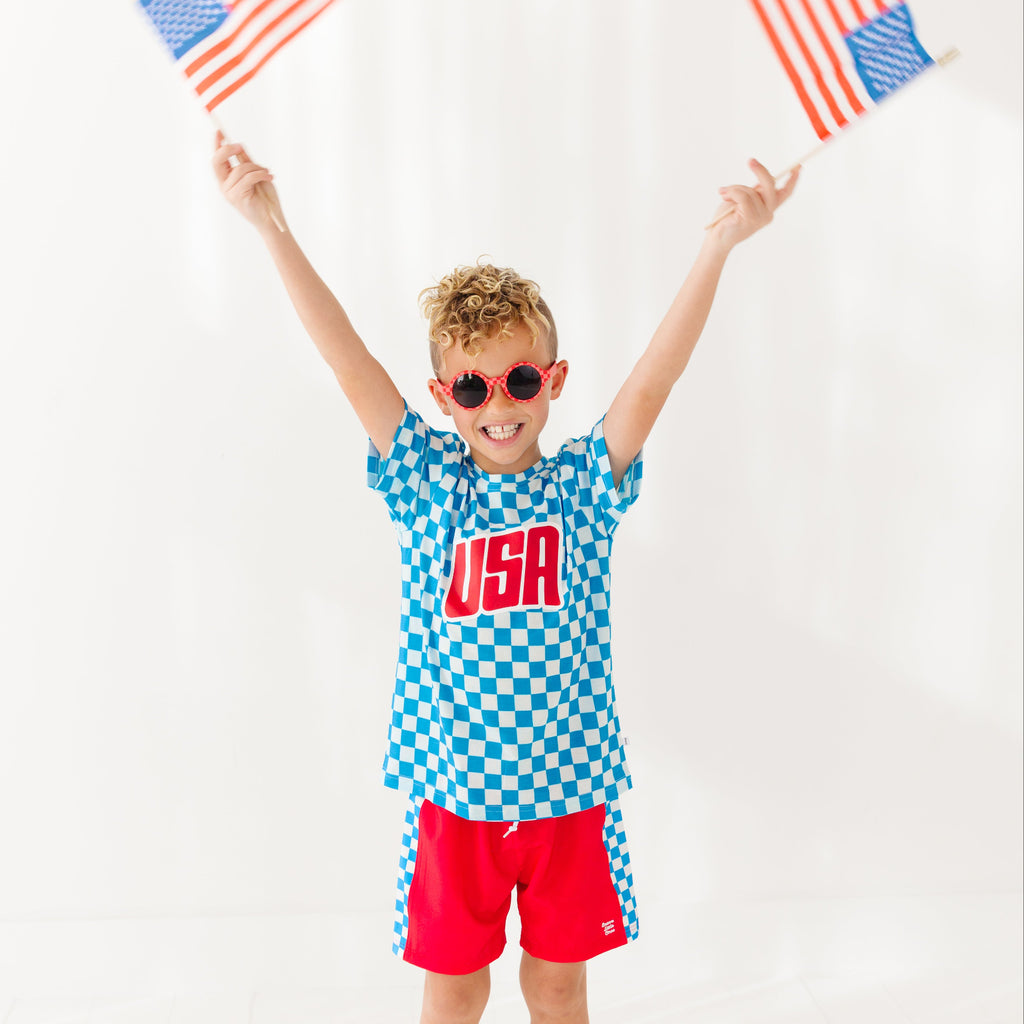Child wearing a blue checkered shirt with 'USA' and red shorts, holding American flags against a white background.