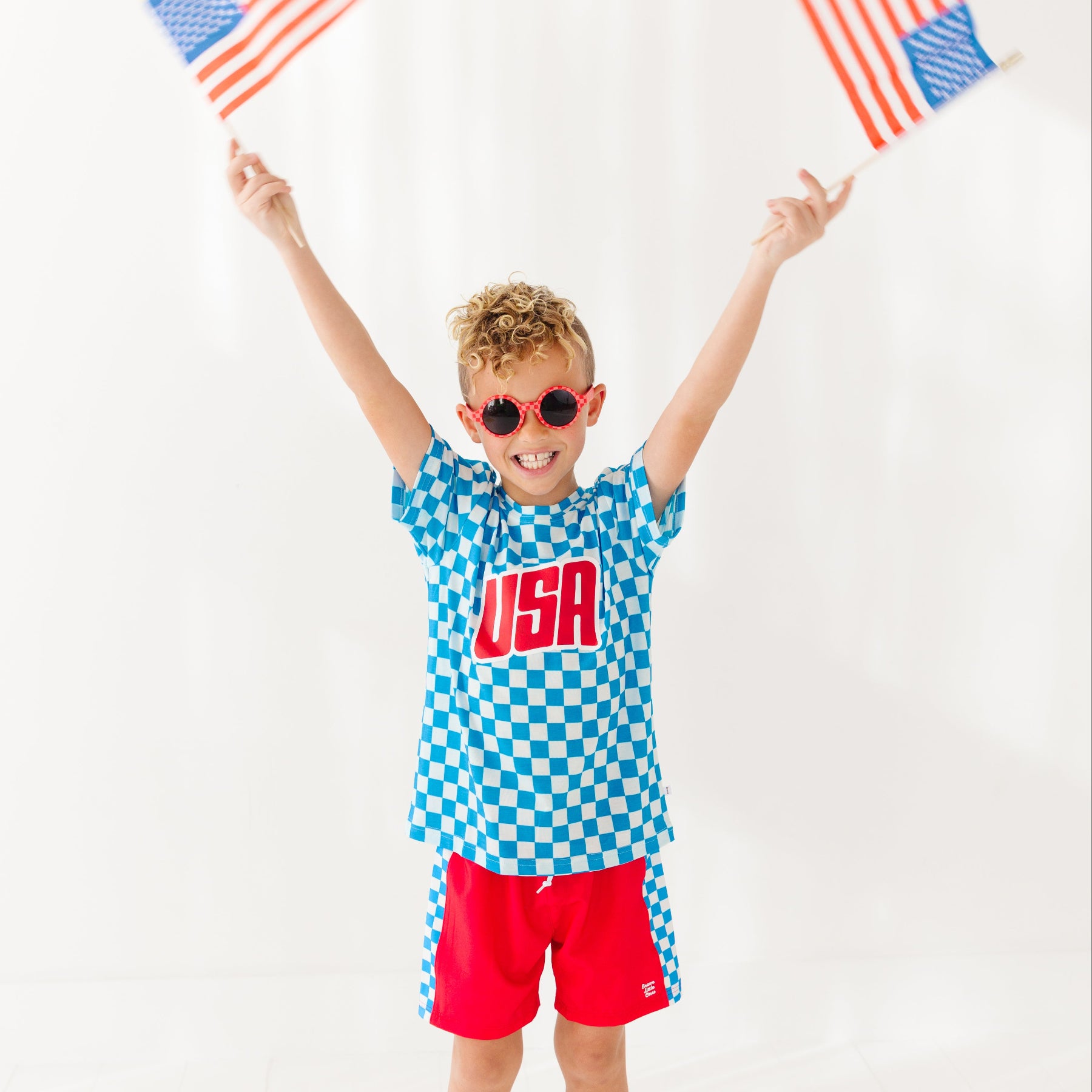 Child wearing a blue checkered shirt with 'USA' and red shorts, holding American flags against a white background.