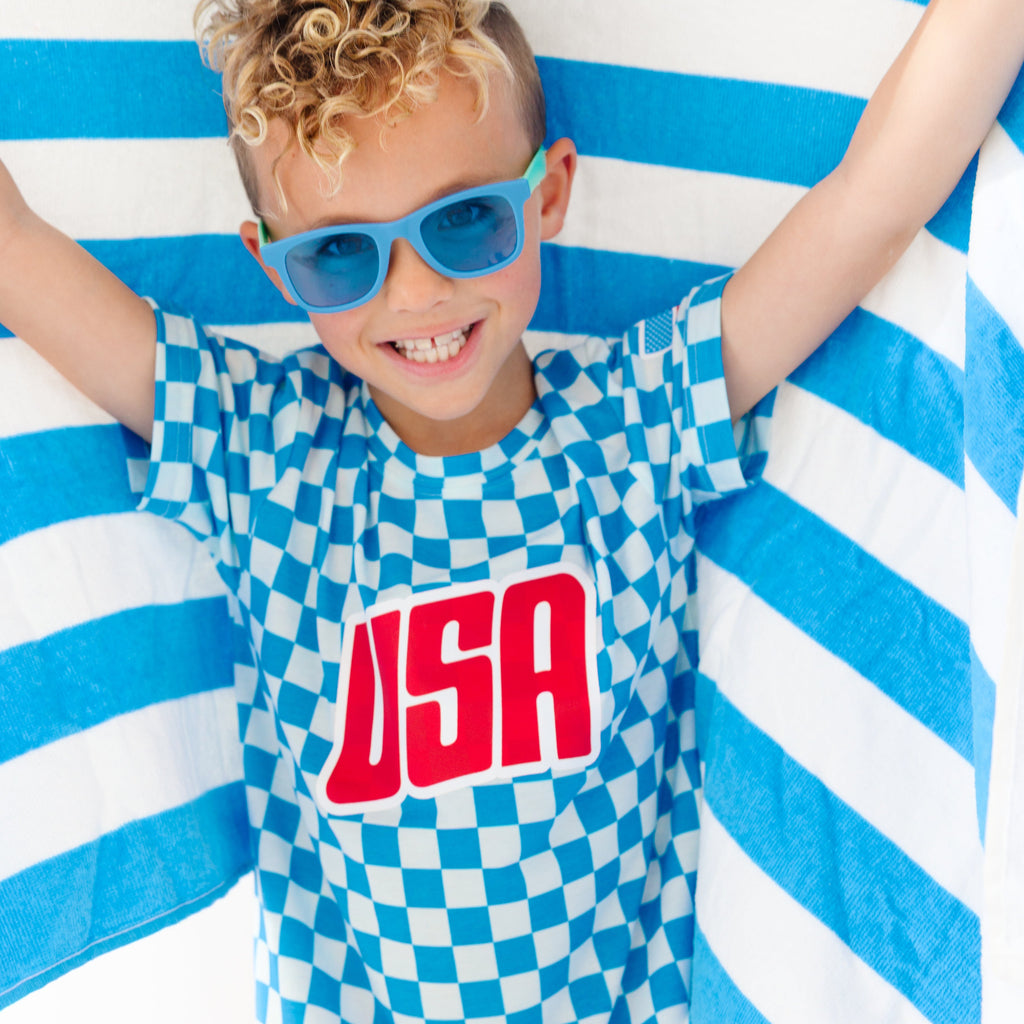 Child wearing a blue and white checkered shirt with 'USA' printed on it, standing against a blue and white striped towel fabric.