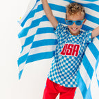Child wearing a blue and white checkered shirt with 'USA' printed on it, standing against a blue and white striped towel fabric.