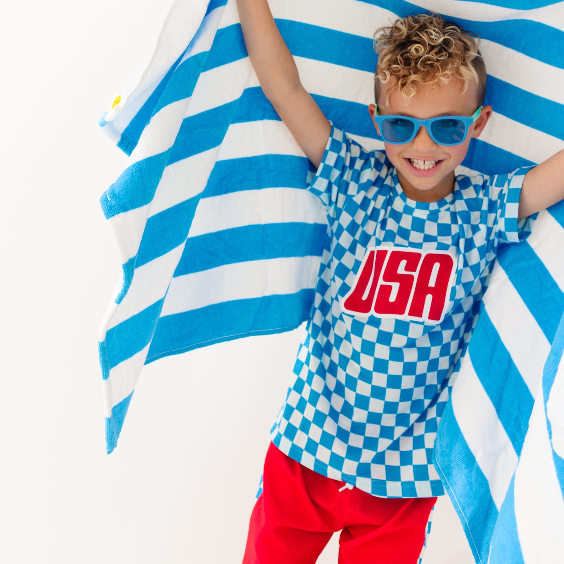 Child wearing a blue and white checkered shirt with 'USA' printed on it, standing against a blue and white striped towel fabric.